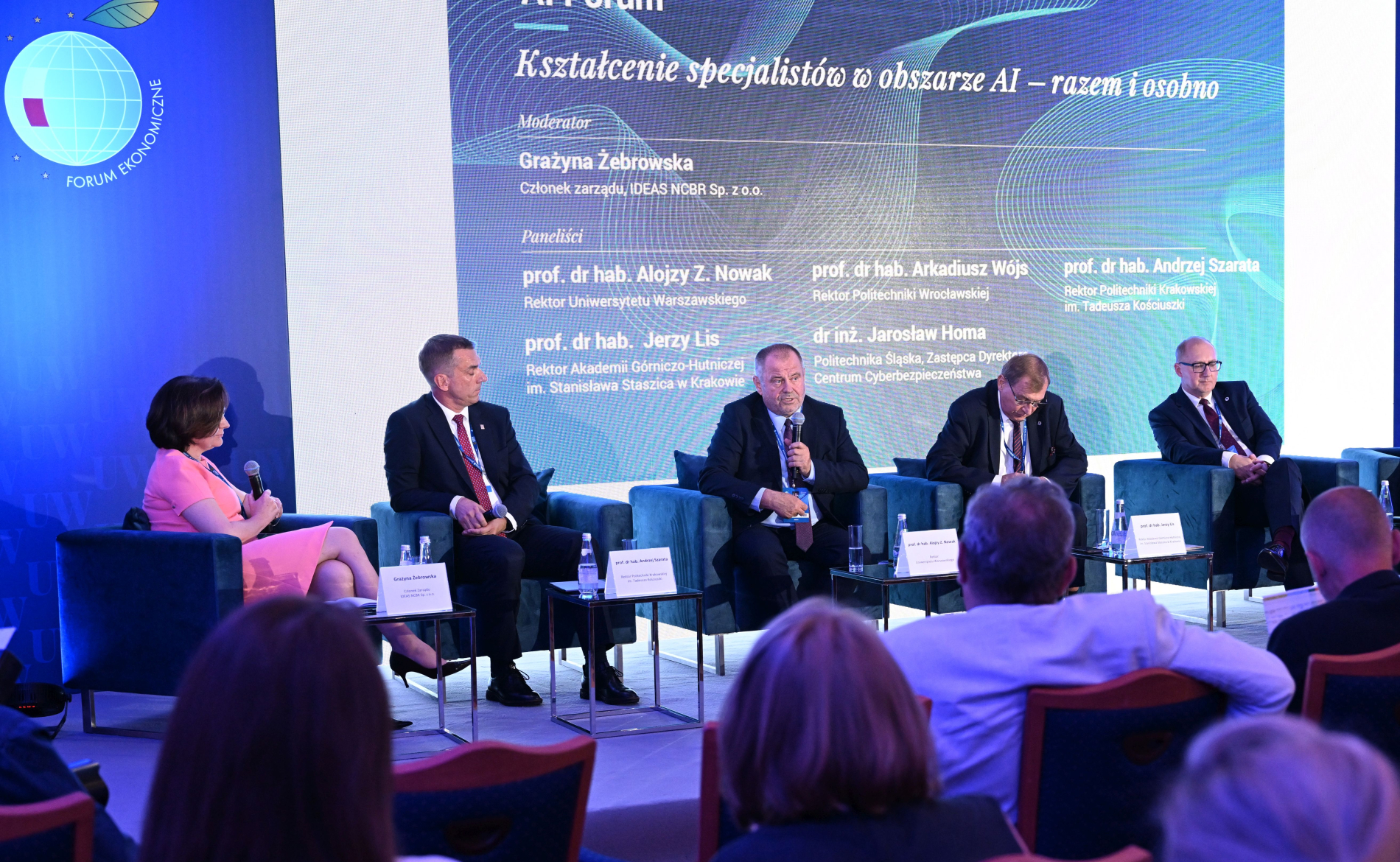 Panel dyskusyjny na Forum Ekonomicznym w Karpaczu. Sześciu prelegentów (w tym jedna kobieta) siedzi na niebieskich fotelach na scenie. Na ekranie w tle widoczny jest tytuł "AI Forum" i lista mówców. W dolnej części zdjęcia widoczne są plecy publiczności.