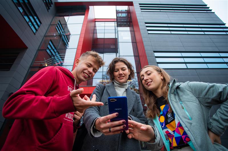 Troje studentów ogląda coś na smartfonie, stojąc na zewnątrz nowoczesnego budynku uczelni z czerwono-czarną fasadą.