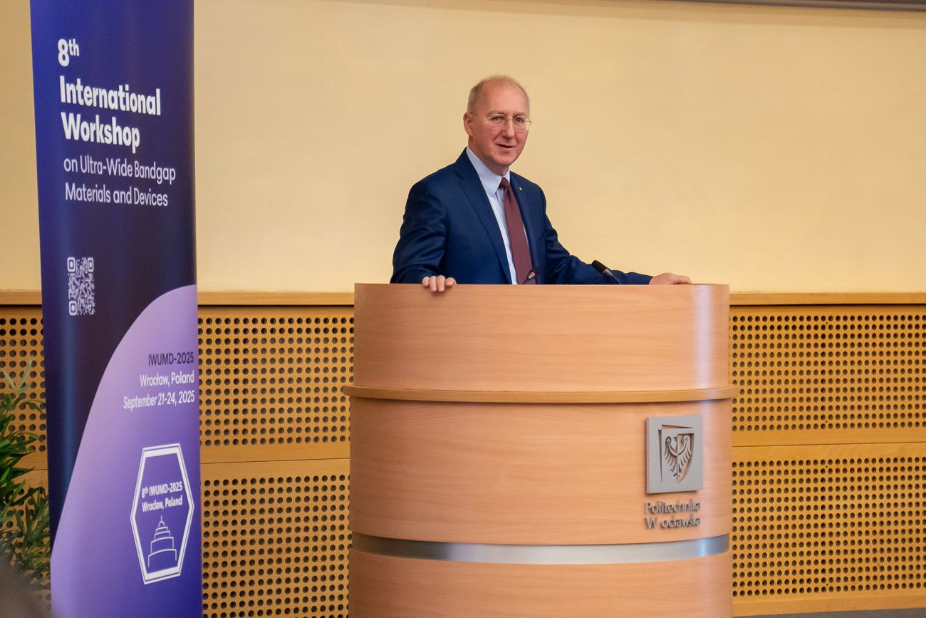 Mężczyzna w garniturze i czerwonym krawacie stoi za drewnianą mównicą. Uśmiecha się i opiera dłonie o boki mównicy. Obok niego widoczny jest pionowy, niebieski baner reklamowy wydarzenia.