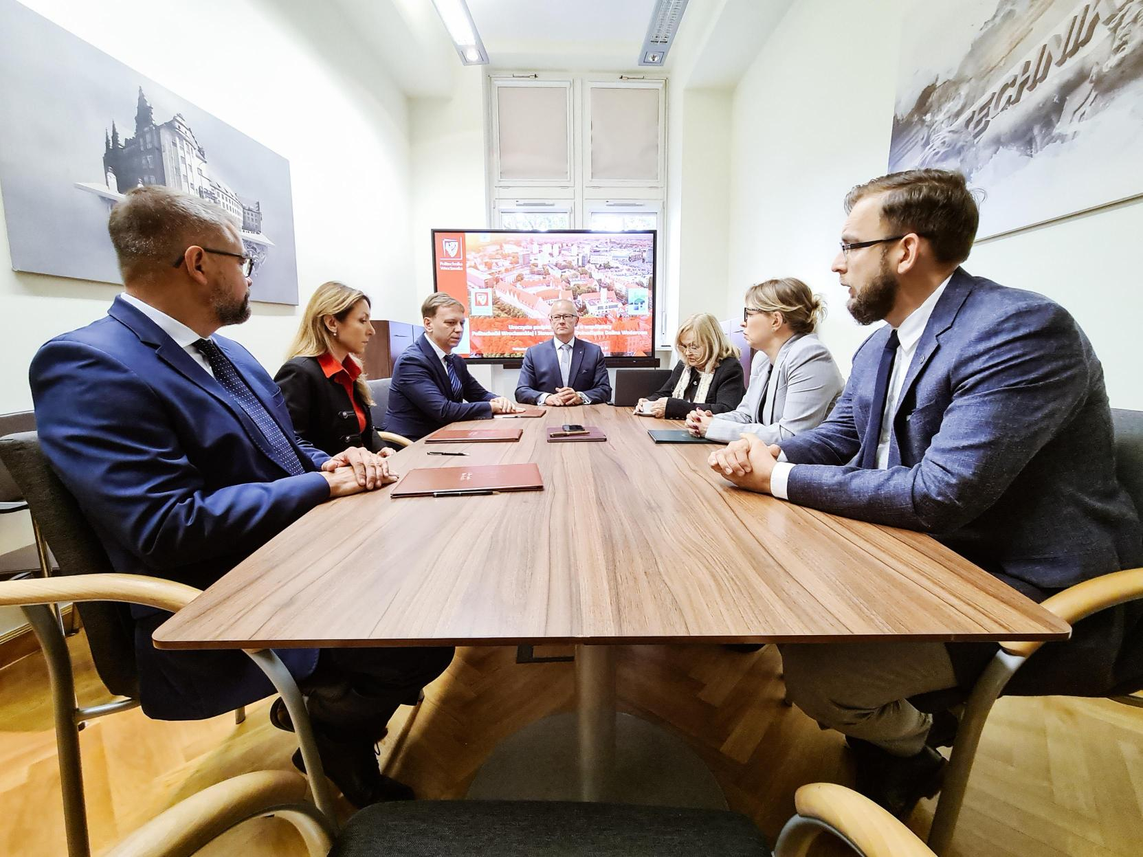 Sześć osób w strojach biznesowych siedzi przy dużym, drewnianym stole konferencyjnym, prowadząc spotkanie.