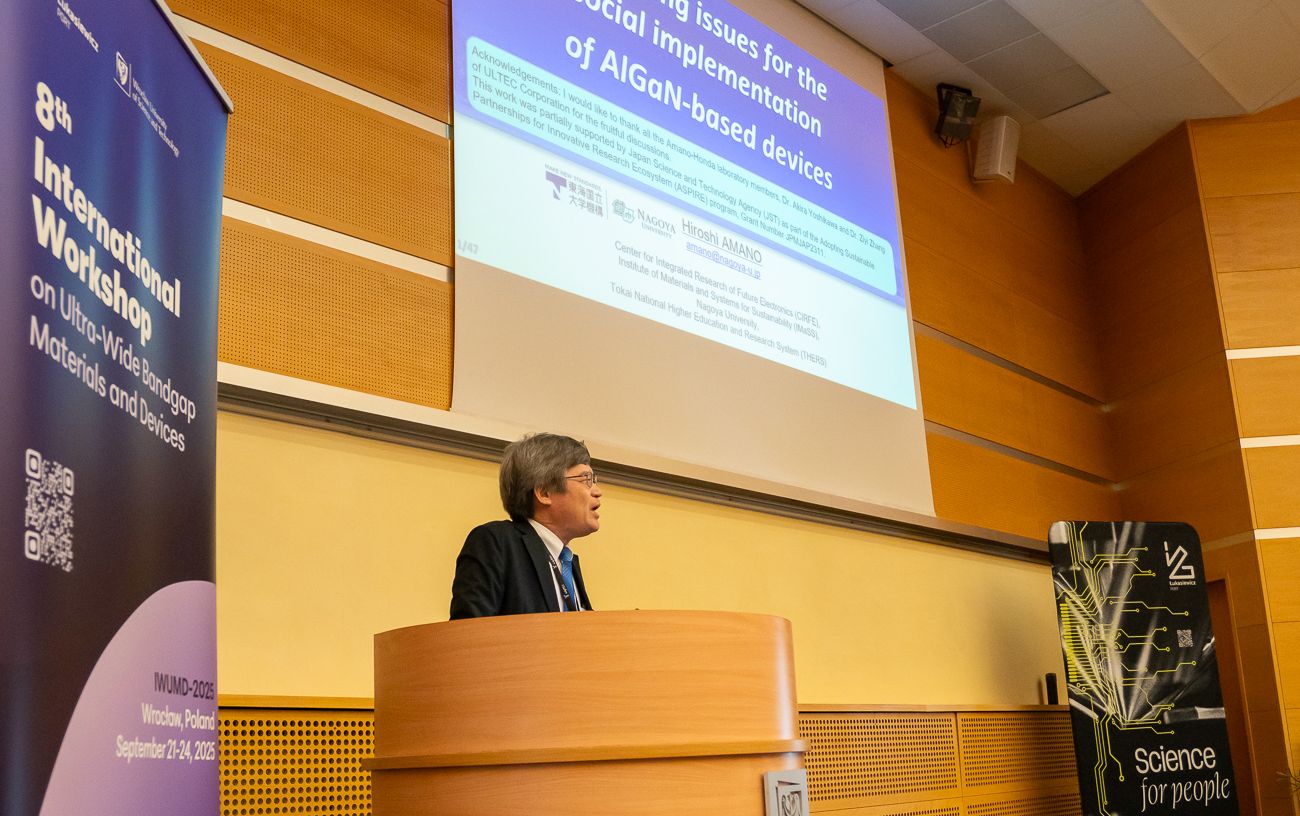 Mężczyzna w garniturze stoi za mównicą, prezentując slajd na ekranie. Na slajdzie widoczne są teksty w języku angielskim oraz logo Politechniki Wrocławskiej i Uniwersytetu w Nagoi. Obok mównicy znajduje się baner warsztatu.