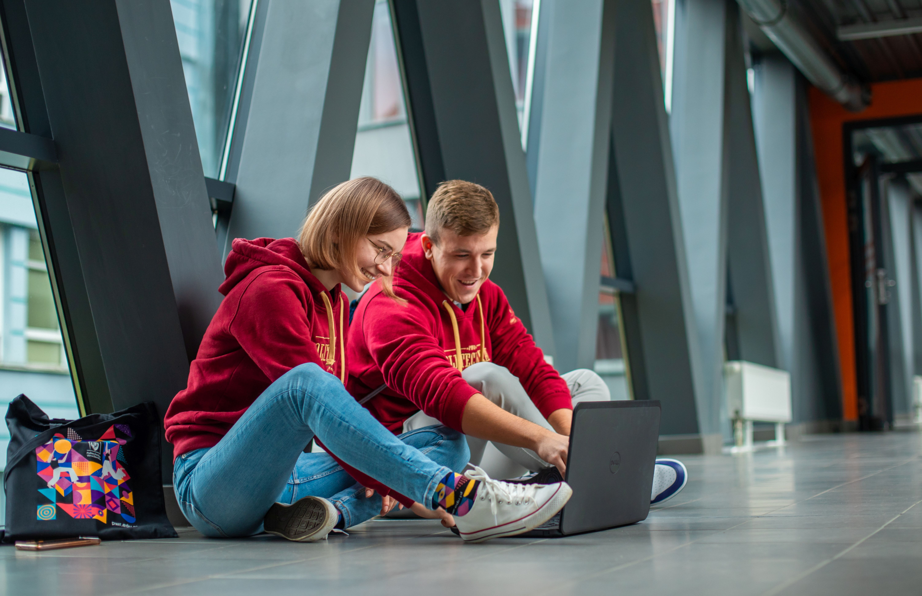 Two people in PWr hoodies are sitting in a hallway, smiling as they look at a laptop.